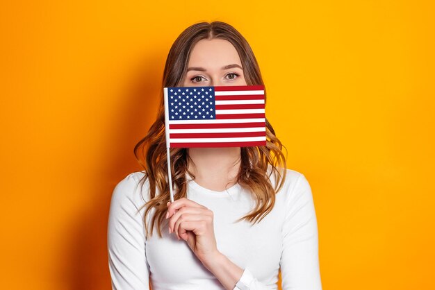 Educação no conceito de América. Jovem caucasiana segura uma pequena bandeira americana isolada de fundo laranja, garota segurando a bandeira dos EUA, 4 de julho dia da independência