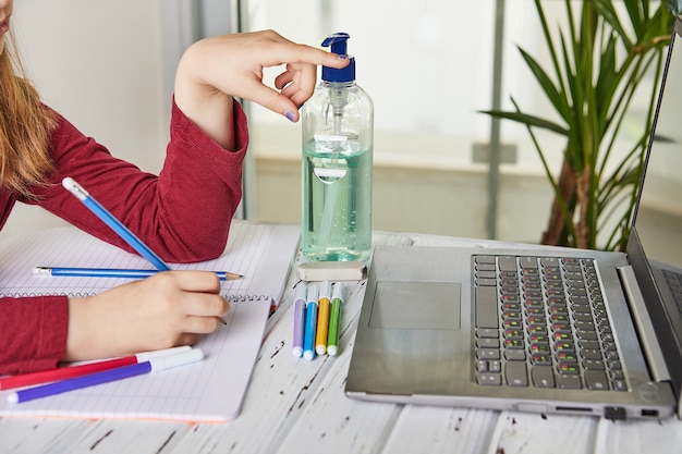 Educação online a distância. colegial estudando em casa com um laptop e fazendo lição de casa da escola. livros de treinamento e canetas de feltro coloridas sobre a mesa, gel com álcool 70%