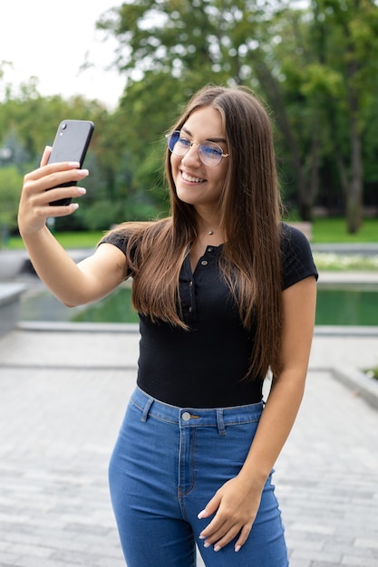 Foto educação online, tecnologia e conceito de internet - uma sorridente menina europeia de cabelos escuros em roupas casuais está em um parque no verão conversando com seus colegas por link de vídeo em sala de aula.