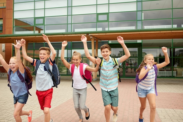 Foto educação primária, amizade, infância e conceito de pessoas - grupo de alunos felizes do ensino primário com mochilas correndo e acenando com as mãos ao ar livre