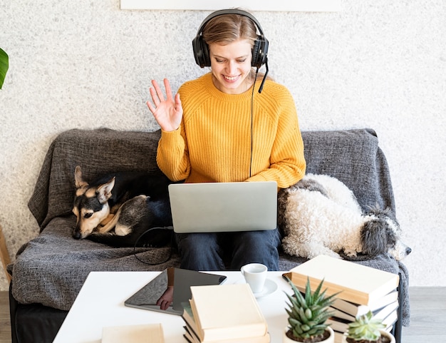 La educación a distancia. Aprendizaje electrónico. Mujer sonriente joven en suéter amarillo y auriculares negros que estudian en línea usando la computadora portátil, sentado en el sofá en casa