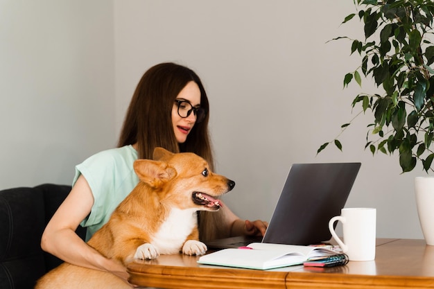 Educación a distancia Chica estudiante con gafas con laptop estudia en línea con su encantador perro Corgi en casa Mujer joven abrazo Welsh Corgi Pembroke Estilo de vida con mascota doméstica