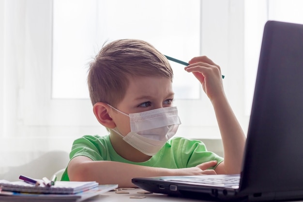 Educación a distancia, educación en línea. Distancia social, autoaislamiento durante la cuarentena. Niños en edad preescolar o escolar en máscara médica estudiando en casa con una computadora portátil, haciendo la tarea para la escuela de desarrollo