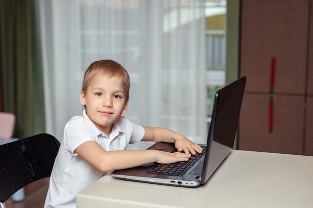 educación a distancia en el hogar para niños durante la cuarentena. Bebé feliz en polo blanco haciendo los deberes usando la computadora portátil sentado en su casa en la cocina