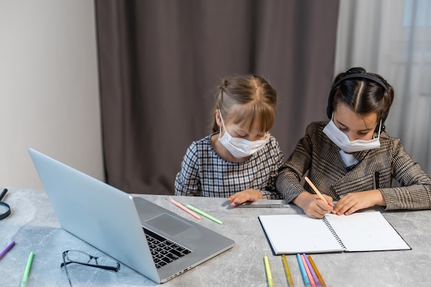 La educación a distancia. Niñas de la escuela en la computadora portátil con máscara estudiando en línea sentado en la mesa en casa