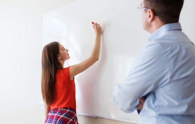 educación, escuela, aprendizaje y concepto de personas - estudiante que escribe algo en una pizarra en blanco y maestra en el aula