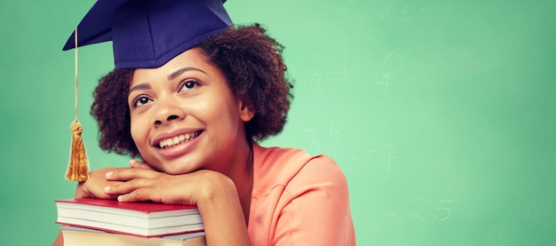 educación, escuela, conocimiento y concepto de la gente - feliz sonriente estudiante afroamericana con gorra de soltero con libros sentados en la mesa y soñando sobre fondo de pizarra verde