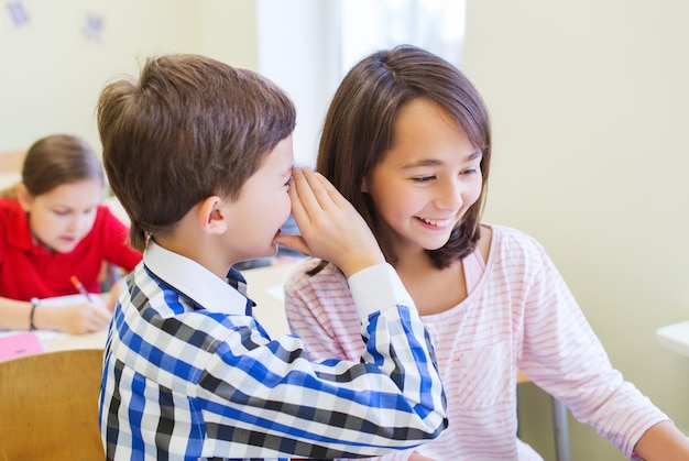 Foto educación, escuela primaria, aprendizaje y concepto de personas - colegial sonriente susurrando secretos al oído de un compañero de clase en el aula