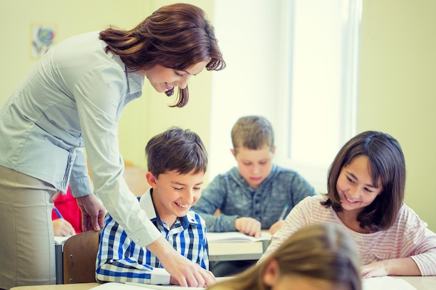 educación, escuela primaria, aprendizaje y concepto de personas - maestro que ayuda a los escolares a escribir pruebas en el aula