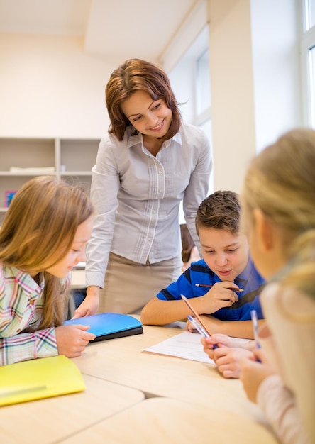 educación, escuela primaria, aprendizaje y concepto de personas - maestro que ayuda a los escolares a escribir pruebas en el aula