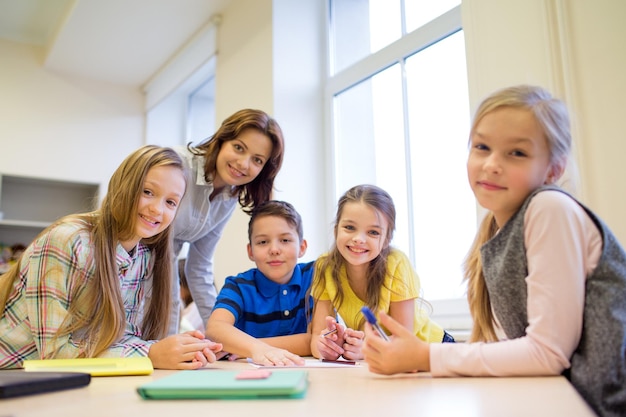 educación, escuela primaria, aprendizaje y concepto de personas - maestro que ayuda a los escolares a escribir pruebas en el aula