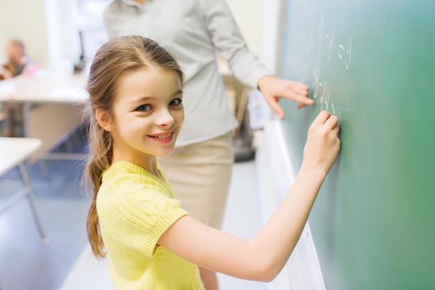 educación, escuela primaria, aprendizaje, matemáticas y concepto de la gente - pequeña colegiala sonriente escribiendo números en una pizarra verde en el aula