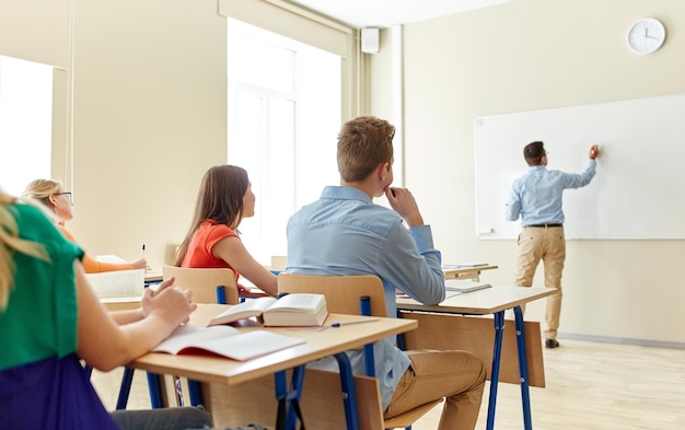 educación, escuela secundaria, aprendizaje, enseñanza y concepto de personas: profesor parado frente a los estudiantes y escribiendo algo en una pizarra blanca en el aula