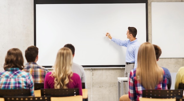 educación, escuela secundaria, trabajo en equipo y concepto de personas: profesor sonriente parado frente a los estudiantes y escribiendo algo en una pizarra blanca en el aula