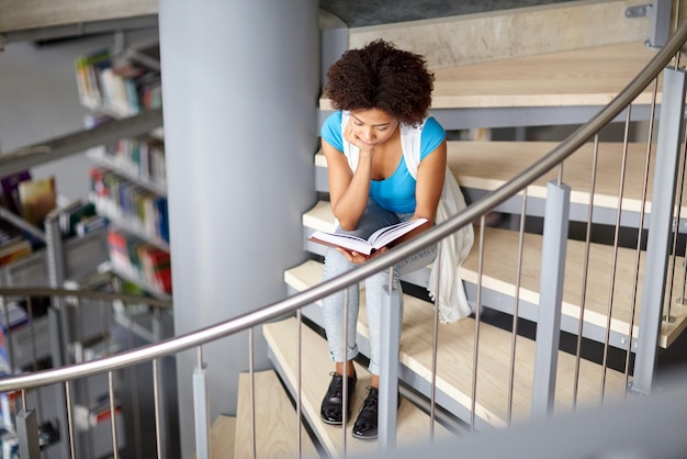 educación, escuela secundaria, universidad, aprendizaje y concepto de la gente - estudiante afroamericana leyendo un libro sentado en las escaleras de la biblioteca
