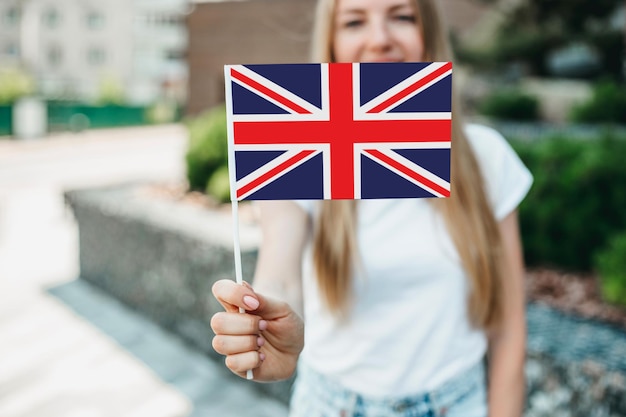 Educación en el extranjero. Chica estudiante se cubre la cara con una pequeña bandera de Inglaterra británica del Reino Unido y se encuentra cerca de la universidad en la calle. Ð¡Ð¾espacio de copia. De cerca