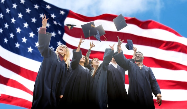 Foto educación, graduación y concepto de la gente - grupo de felices estudiantes internacionales vestidos de soltero lanzando mortarboards sobre el fondo de la bandera estadounidense