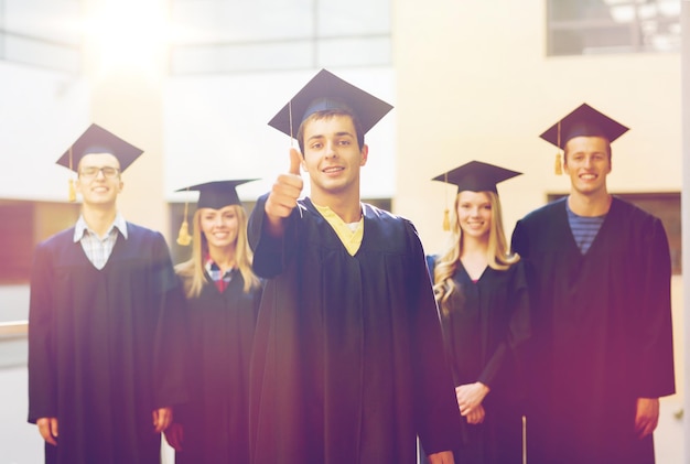 educación, graduación, gesto y concepto de personas - grupo de estudiantes sonrientes en mortarboards y batas mostrando los pulgares hacia arriba al aire libre