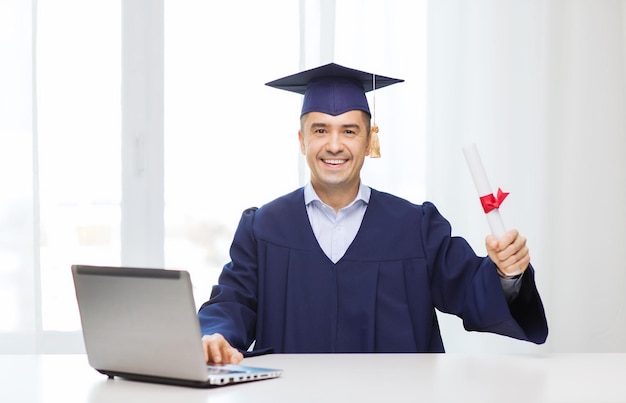 educación, graduación, negocios, tecnología y concepto de personas - estudiante adulto feliz en birrete con diploma y computadora portátil sentado en la mesa en casa u oficina