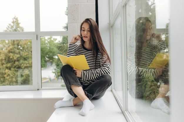 Foto educación en el hogar una adolescente una estudiante se sienta en un alféizar de la ventana escribe la tarea preparación de aprendizaje a distancia para los exámenes enfoque selectivo suave