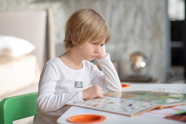 Educación en el hogar durante la pandemia. Un niño aburrido leyendo un libro en casa.