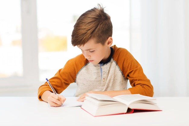 educación, infancia, gente, deberes y concepto escolar - estudiante con libro escribiendo en el cuaderno en casa