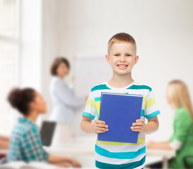 educación, infancia, trabajo en equipo y concepto escolar - niño estudiante sonriente con libro azul sobre un grupo de estudiantes en el aula