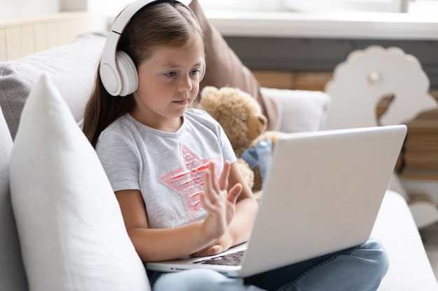 Educación en línea de niños. Dulce niña con auriculares mirando portátil de conferencia de profesor de video lección sentado en el sofá en casa.