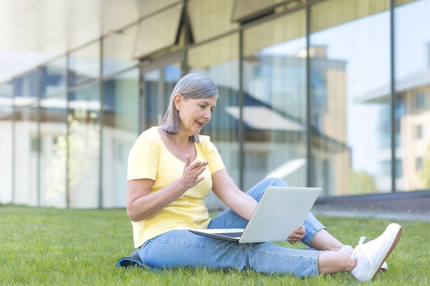 Educación en línea Retrato de una anciana hermosa sentada en el campus en el césped con una computadora portátil y una bicicleta Hablando en una videollamada en una computadora portátil agitando los brazos sonriendo