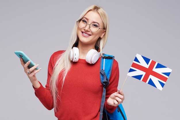 Educación en línea, traductor de idiomas extranjeros, inglés, estudiante - mujer rubia sonriente en auriculares con teléfono móvil y bandera británica. Espacio gris, aprendizaje a distancia