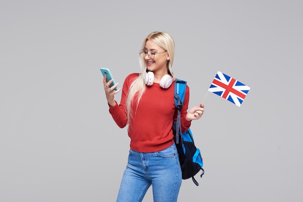 Educación en línea, traductor de idiomas extranjeros, inglés, estudiante - mujer rubia sonriente en auriculares con teléfono móvil y bandera británica. Espacio gris, aprendizaje a distancia