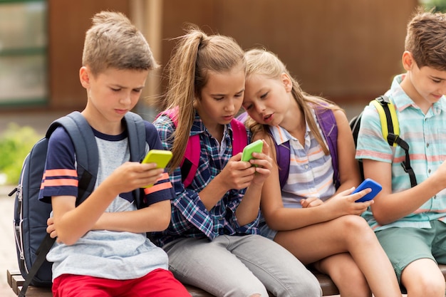 educación primaria, amistad, infancia, tecnología y concepto de personas - grupo de felices estudiantes de primaria con smartphones y mochilas sentados en un banco al aire libre