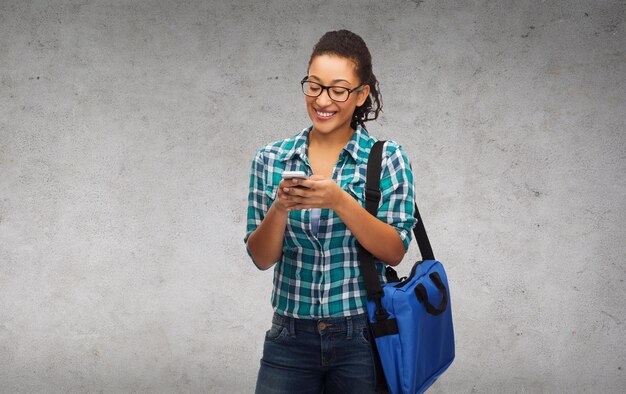 educación, tecnología y concepto de personas - estudiante afroamericana sonriente en anteojos con teléfono inteligente y bolsa
