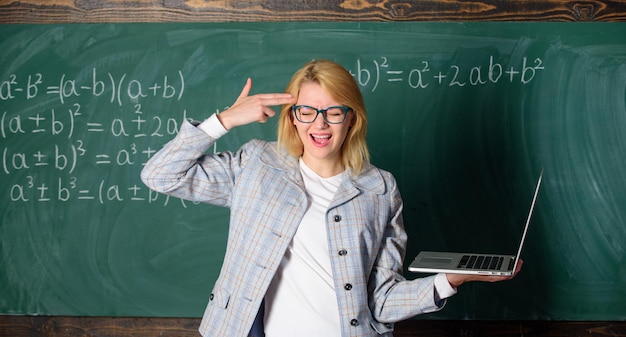 Educadora inteligente inteligente com laptop moderno segura gesto de arma em seu fundo de quadro-negro. Professor cansado irritado por seu trabalho. Mulher cansada ou irritada detém laptop. Conceito de reformas educacionais.