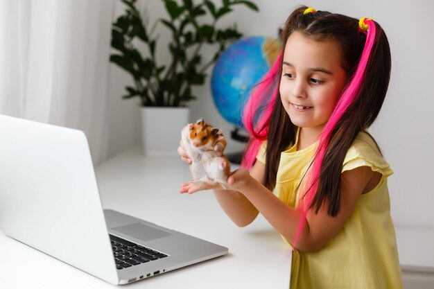 Educar en casa. La muchacha del niño hace la tarea con el hámster del animal doméstico. Hámster de jengibre divertido sentado en la mesa donde el niño está escribiendo. De vuelta a la escuela.