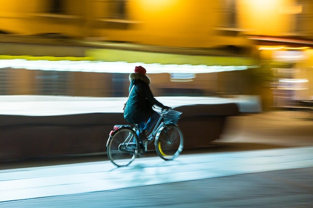 Efeito panorâmico de uma senhora que está andando de bicicleta à noite entre a iluminação da cidade Ainda assunto e fundo em movimento