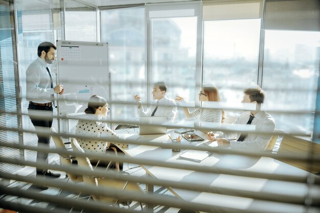 Effektives Brainstorming. Angenehme junge Kollegen, die am Tisch sitzen und ihre Ideen bezüglich des Projekts teilen, während ihr Chef in der Nähe des Whiteboards steht und ihnen zuhört