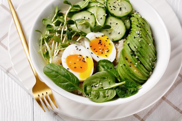 Egg Topped Quinoa Bowl mit Sonnenblumen-Mikrogrün, Gurke, Avocado, Babyspinat in einer weißen Schüssel auf einem weißen Holztisch, türkische Küche