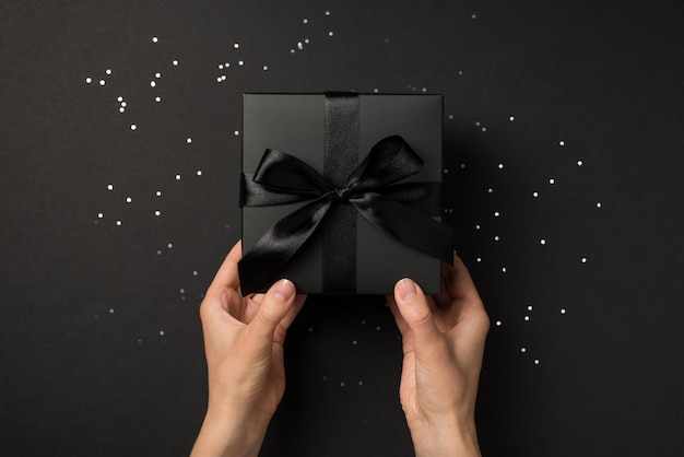 Ego-Draufsichtfoto von Händen, die eine schwarze Geschenkbox mit schwarzer Schleife über Pailletten auf isoliertem schwarzem Hintergrund halten