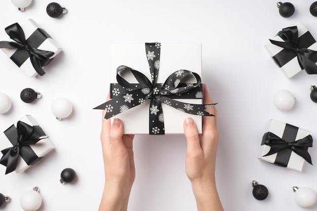 Ego-Draufsichtfoto von weiblichen Händen, die eine weiße Geschenkbox mit schwarzer Satinbandschleife über weißen und schwarzen Weihnachtsdekorationen, Geschenken und Bällen auf isoliertem weißem Hintergrund geben
