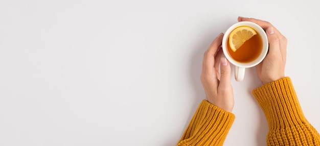 Ego-Draufsichtfoto von weiblichen Händen im gelben Pullover, die eine weiße Tasse Tee mit Zitronenscheibe auf isoliertem weißem Hintergrund mit Copyspace berühren