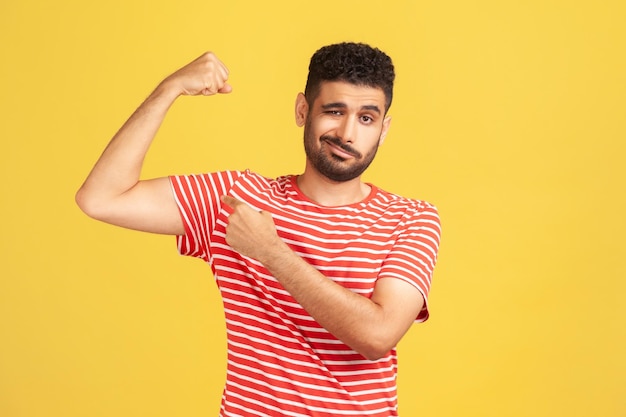 Egoistischer, selbstbewusster Mann mit Bart in gestreiftem T-Shirt, der mit dem Finger auf den Armbizeps zeigt und seine Stärke und sein unabhängiges Führungskonzept zeigt Studioaufnahme im Innenbereich isoliert auf gelbem Hintergrund