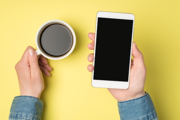 Egoperspektive Foto von Frauenhänden, die eine Tasse Kaffee und ein Smartphone auf isoliertem gelbem Hintergrund mit Copyspace halten