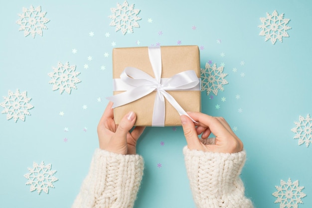Egoperspektive Foto von Händen in weißem Pullover beim Auspacken einer Geschenkbox aus Bastelpapier mit weißer Schleife über großen dekorativen Schneeflocken und glänzendem Konfetti auf isoliertem pastellblauem Hintergrund