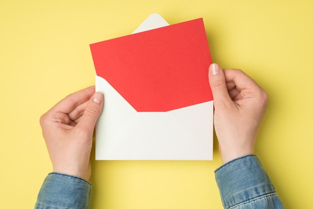 Egoperspektive Foto von weiblichen Händen, die einen offenen weißen Umschlag mit roter Papierkarte auf isoliertem gelbem Hintergrund mit Leerzeichen halten