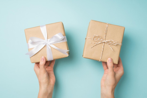 Egoperspektive Foto von weiblichen Händen, die zwei Kraftpapier-Geschenkboxen mit Bindfäden und weißer Satinbandschleife auf isoliertem pastellblauem Hintergrund halten