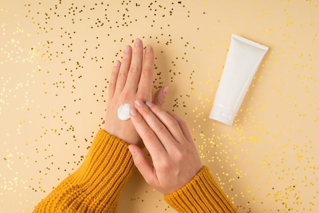 Egoperspektive Foto von weiblichen Händen in gelbem Pullover, die cremeweiße Cremeröhrchen und verstreute glänzende goldene Pailletten auf isoliertem pastellorangefarbenem Hintergrund mit Leerzeichen auftragen