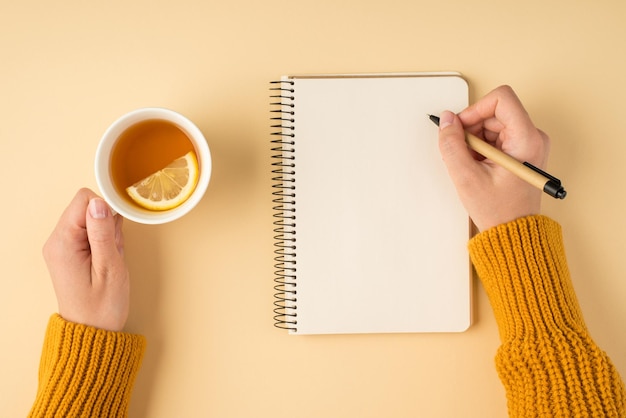 Egoperspektive Foto von weiblichen Händen in gelbem Pullover, die in Spiralplaner schreiben und eine weiße Tasse Tee mit Zitronenscheibe auf isoliertem pastellorangefarbenem Hintergrund mit Kopierraum halten