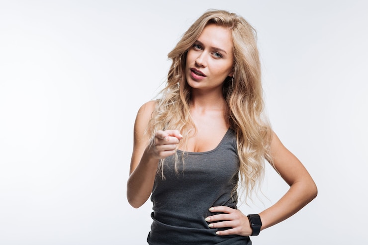 Foto eh, tú. hermosa joven rubia en una camiseta sin mangas gris con una mano en la cintura y apuntando mientras está de pie aislado sobre fondo gris