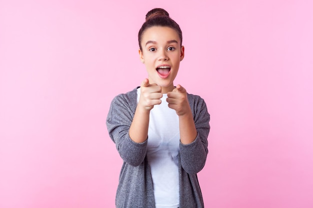 Eh, tú. Retrato de una adolescente morena sorprendida con peinado de moño con ropa informal apuntando a la cámara, de pie con la boca abierta, expresión asombrada. foto de estudio, fondo rosa